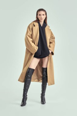 Bej keçeli ceket ve yüksek topuklu siyah deri çizme giyen şık, kendine güvenen bir kadının tam boy portresi. Beyaz stüdyo geçmişi. Kadın modası.