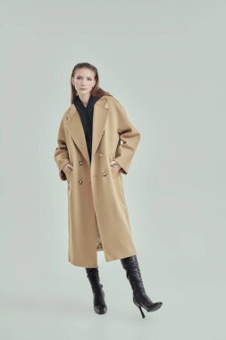 Bej keçeli ceket ve yüksek topuklu siyah deri çizme giyen şık, kendine güvenen bir kadının tam boy portresi. Beyaz stüdyo geçmişi. Kadın modası.