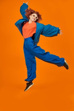 Mavi ve turuncu giysiler içinde atlayan parlak kırmızı kıvırcık saçlı, neşeli model kızın tam boy portresi. Parlak renkler. Stüdyo turuncu arka plan. Yaz modası. 