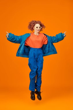 Mavi ve turuncu giysiler içinde atlayan parlak kırmızı kıvırcık saçlı, neşeli model kızın tam boy portresi. Parlak renkler. Stüdyo turuncu arka plan. Yaz modası. 
