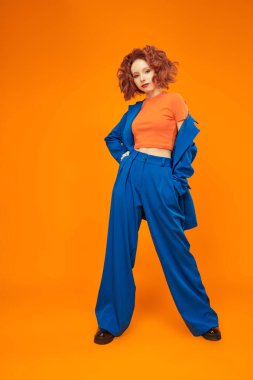 Parlak kırmızı kıvırcık saçlı, mavi ve turuncu elbiseli çekici, neşeli model kızın tam boy portresi. Parlak renkler. Stüdyo turuncu arka plan. Yaz modası. 