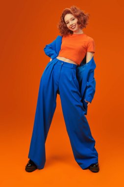 Parlak kırmızı kıvırcık saçlı, mavi ve turuncu elbiseli çekici, neşeli model kızın tam boy portresi. Parlak renkler. Stüdyo turuncu arka plan. Yaz modası. 