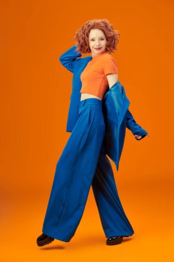 Parlak kırmızı kıvırcık saçlı, mavi ve turuncu elbiseli çekici, neşeli model kızın tam boy portresi. Parlak renkler. Stüdyo turuncu arka plan. Yaz modası. 