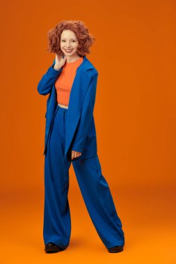 Parlak kırmızı kıvırcık saçlı, mavi ve turuncu elbiseli çekici, neşeli model kızın tam boy portresi. Parlak renkler. Stüdyo turuncu arka plan. Yaz modası. 