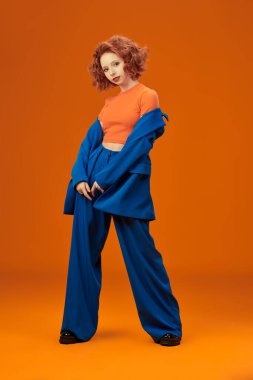 Parlak renkler. Parlak kırmızı kıvırcık saçlı, mavi ve turuncu elbiseli çekici, neşeli model kızın tam boy portresi. Stüdyo turuncu arka plan. Yaz modası. 
