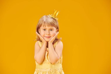 Sarı saçlı, sarı elbiseli ve altın tacı olan gülümseyen küçük bir prensesin portresi. Sarı stüdyo geçmişi. Mutlu çocukluk, mutlu çocuk duyguları.