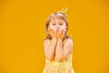 Sarı saçlı, sarı elbiseli ve altın tacı olan gülümseyen küçük bir prensesin portresi. Sarı stüdyo geçmişi. Mutlu çocukluk, mutlu çocuk duyguları.