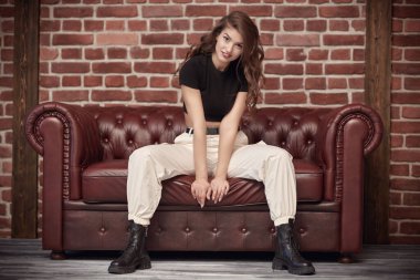 Çekici, kendine güvenen, uzun kıvırcık saçlı, siyah bluzlu, spor pantolonlu, siyah çizmeli, deri koltukta poz veren genç bir kadın. Güzellik ve moda. Kendine güvenen yaşam tarzı.