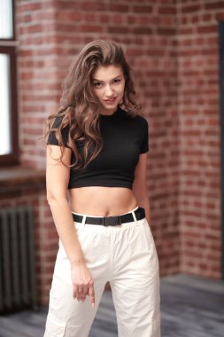 Kendine güvenen bir yaşam tarzı. Çekici, kendine güvenen, uzun kıvırcık saçlı, siyah bluzlu ve beyaz pantolonlu bir çatı katında poz veren genç bir kadın. Güzellik ve moda.