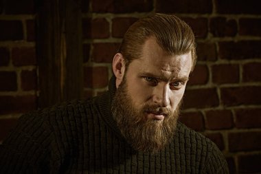 Cesaret ve güç. Büyük sakallı bir adam portresi örgü kazak, grunge duvara karşı duran ve sakince ve emin bir şekilde ileriye bakıyor.