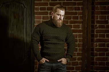 Courage and strength of men. A portrait of a large bearded man in a knitted sweater, who stands against the backdrop of a brick wall and calmly and confidently looks ahead. clipart