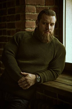 Tuğla bir duvarın arkasında sakince ve düşünceli bir şekilde duran örgü örülü süveter giyen yakışıklı bir sakallı adamın portresi..