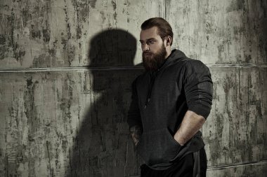 Erkeklik ve fiziksel güç kavramı. Siyah kapüşonlu, iri yarı, sakallı bir adamın portresi. Arka planda beton bir duvar var..