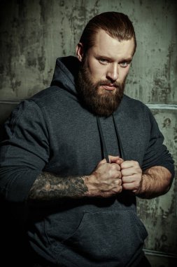 Shot of a muscular brutal bearded man in a black hoodie against a concrete wall. The concept of masculinity and physical strength.  clipart