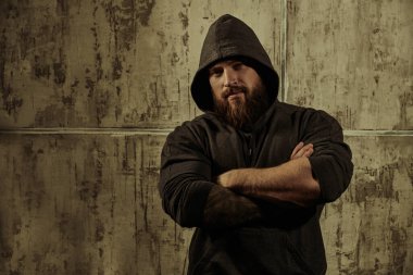 Siyah kapüşonlu, beton duvara dayanmış kaslı, kaba sakallı bir adamın portresi. Erkeklik ve fiziksel güç kavramı. Metin için yer.