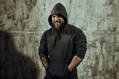 Erkeklik ve fiziksel güç kavramı. Siyah kapüşonlu, beton duvara dayanmış kaslı, kaba sakallı bir adamın portresi. Metin için yer.