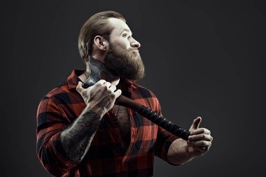 Baltayla tıraş olan, ekose gömlekli, vahşi sakallı bir adamın portresi. Koyu gri stüdyo geçmişi. Erkek güzelliği ve erkeklik. Berber dükkanı reklamı.