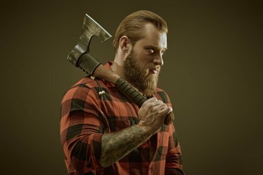 Erkek güzelliği ve erkeklik. Elinde baltası olan, ekose gömlekli, vahşi sakallı bir adamın portresi. Koyu kahverengi stüdyo geçmişi. Berber dükkanı reklamı.