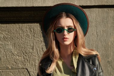 Boho ve hipster tarzı. Şık güneş gözlüklü, şapkalı ve deri ceketli güzel hippi kız güneşe bakan bir caddede duruyor. İnsanlar ve yaşam tarzı. Boşluğu kopyala.