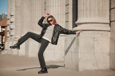 Uzun menzilli çekim. Siyah deri ceketli, siyah kot pantolonlu ve güneş gözlüklü modern bir kadın. Deri kıyafet modası. Çekici bir genç kadın. Moda ve gençlik tarzı. Boşluğu kopyala.