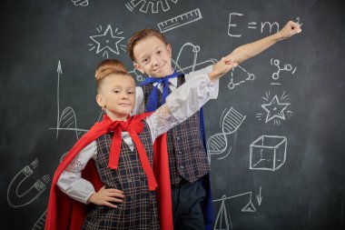 Pelerinli şirin çocuklar süper kahraman pozu veriyor. Eğitim ve erken gelişim. Küçük Bilim adamları. Siyah bir karatahtanın arka planı.