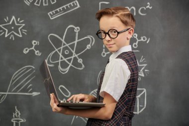 Eğitim. Okul üniformalı ve gözlüklü akıllı bir çocuğun portresi. Kameraya ciddi ciddi dizüstü bilgisayarla bakıyor. Bilimsel çizimleri olan siyah bir kara tahta. Genç bilim adamları.
