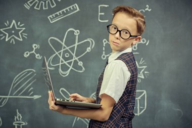Eğitim. Okul üniformalı ve gözlüklü akıllı bir çocuğun portresi. Kameraya ciddi ciddi dizüstü bilgisayarla bakıyor. Bilimsel çizimleri olan siyah bir kara tahta. Genç bilim adamları.