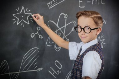 Okul üniformalı ve kameraya ciddi ciddi bakan zeki bir çocuğun portresi. Bilimsel çizimleri olan siyah bir kara tahta. Eğitim. Genç bilim adamları.