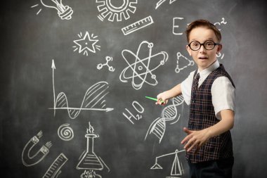 Genç bilim adamı. Okul üniformalı ve kameraya ciddi ciddi bakan zeki bir çocuğun portresi. Bilimsel çizimleri olan siyah bir kara tahta. Eğitim, öğretim.