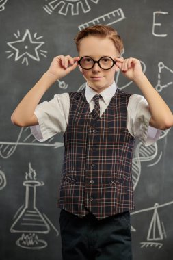 Okul üniformalı ve kameraya ciddi ciddi bakan zeki bir çocuğun portresi. Bilimsel çizimleri olan siyah bir kara tahta. Eğitim. Genç bilim adamları.