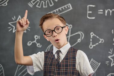 Okul üniformalı, gözlüklü ve kameraya ciddi ciddi bakan zeki bir çocuğun portresi. Bilimsel çizimleri olan siyah bir kara tahta. Eğitim. Genç bilim adamları.