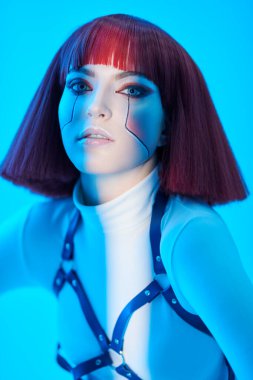 Parlak kırmızı makyajlı ve şık kırmızı saçlı seksi siber punk kız beyaz yüksek teknoloji kıyafetlerle mavi ışıkta beyaz arka planda poz veriyor. Sanal gerçeklik, oyun. Siber Moda..