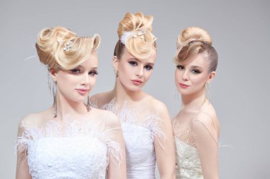Düğün makyajı ve saç stili. Parlak makyajlı ve zarif saç stili olan beyaz gelinlikli üç güzel sarışın kız. Açık gri arkaplanda stüdyo portresi. 