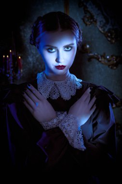 Beyaz yakalı, düzgün siyah elbiseli esmer bir kadın kasvetli gotik bir şatoda gece ayakta duruyor ve kameraya pek dostça görünmüyor. Gotik kara komedi. Addams Ailesi. 