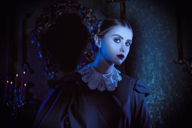 Dost canlısı olmayan siyah elbiseli, beyaz yakalı esmer bir kadın karanlık bir gotik şatoda duruyor ve kameraya pek dostça görünmüyor. Gotik kara komedi. Addams Ailesi. 