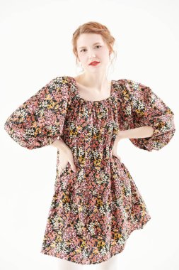 Çiçekli bir elbiseyle beyaz bir duvara poz veren güzel bir kızıl saçlı kızın stüdyo portresi. Romantik kadın tarzı. Bahar ve yaz modası, güzellik.