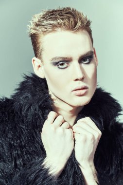 Makyaj, saç stili, makyaj malzemeleri. Kısa saç kesimi ve siyah dumanlı gözleriyle stüdyoda siyah kürklü poz veren şık bir gencin portresi. Moda ve erkek güzelliği. 