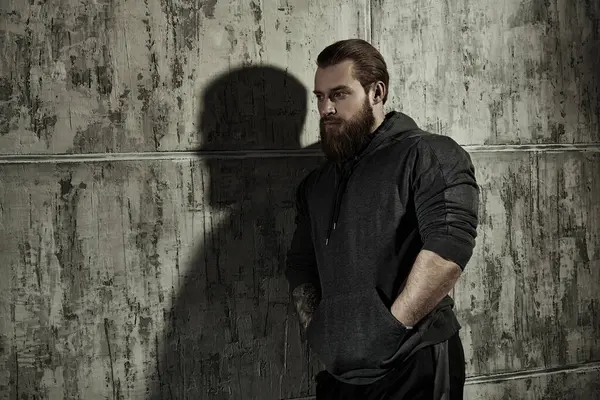 Erkeklik ve fiziksel güç kavramı. Siyah kapüşonlu, iri yarı, sakallı bir adamın portresi. Arka planda beton bir duvar var..