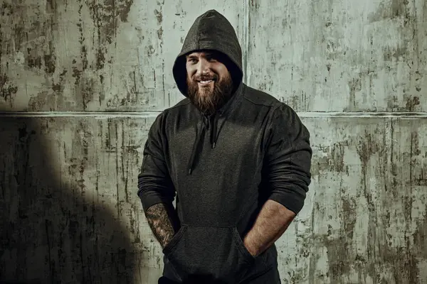 Erkeklik ve fiziksel güç kavramı. Siyah kapüşonlu, beton duvara dayanmış kaslı, kaba sakallı bir adamın portresi. Metin için yer.