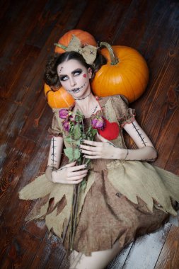 Voodoo tales. A beautiful and dangerous young woman dressed as a voodoo doll lies on a wooden floor, leaning on pumpkins and holding dried roses in her hands. Halloween. clipart