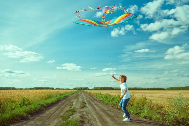 Açık bir alanda güneşli bir günde açık renkli bir uçurtma uçuran açık renk elbiseli, sarışın bir çocuğun tam boy portresi. Yaz zamanı. Çocuk oyunları. Mutlu bir çocukluk. Boşluğu kopyala.
