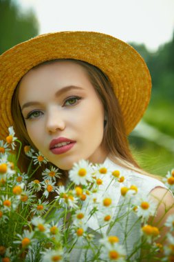 Yazın hasır şapkalı, kahverengi saçlı romantik bir kız bir buket taze papatyayla bir yaz günü dışarıda duruyor. Yaz güzelliği, makyaj ve çiçekler..