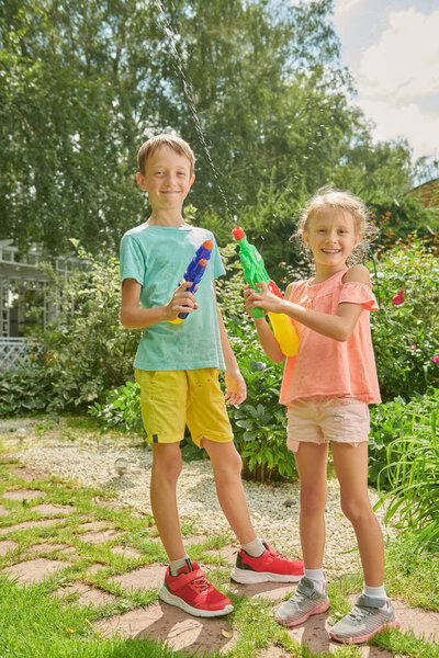 Двое веселых детей играют с водяными пистолетами в саду в летний день. Летняя радость и детские игры. 