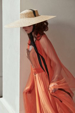 Zarif, çekici bir bayan, güzel, turuncu bir elbise ve geniş kenarlı bir şapkayla yaz şehrinde yürüyor. Yaz modası.