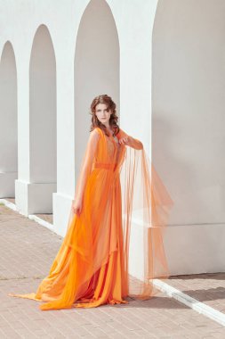 Zarif turuncu elbisesi ve şifonlu atkısı olan güzel genç bir bayan beyaz şehir duvarlarının arkasında duruyor. Romantik yaz tarzı.