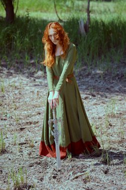 Muhteşem uzun kızıl saçlı, tarihi bir Kelt elbisesi olan genç ve güzel bir kadın kılıçlı bir ormanda duruyor. Orta Çağ. Fantezi. Tarihi yeniden yapılandırma.