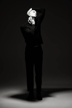 Siyah elbiseli genç bir adamın sanat portresi. Karanlıkta duran ve yüzünü aydınlatan bir ışık huzmesi. Siyah arkaplana karşı tam boy siyah beyaz portre. Psikoloji. Tiyatro.