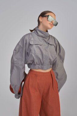 Yüksek moda. Tarz sahibi gri bir ceketle poz veren güzel bir kız modelin portresi, geniş kahverengi pantolon ve gri bir stüdyo arka planında aynalı güneş gözlüğü. Gelecekçi Moda Biçimi. 