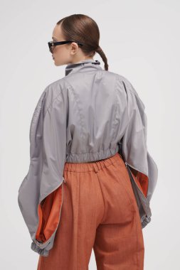 Gri ceketli, geniş kahverengi pantolonlu ve gri arka planda aynalı güneş gözlüklü güzel bir mankenin portresi. Fütürist moda tarzı. Yüksek moda.