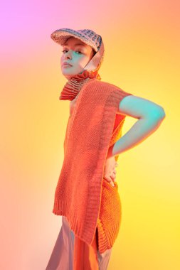 Yarı sezonluk özel tasarım koleksiyonu. Örgü giyim tarzı. Geniş paçalı pançoyla poz veren şık bir manken, atölyede örgü ören panço ve karışık renkli ışıkta örgü ören bir şapka. Parlak renkler.